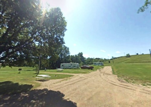 A sunny outdoor scene featuring a gravel road, grassy areas, and parked RVs under a large tree.