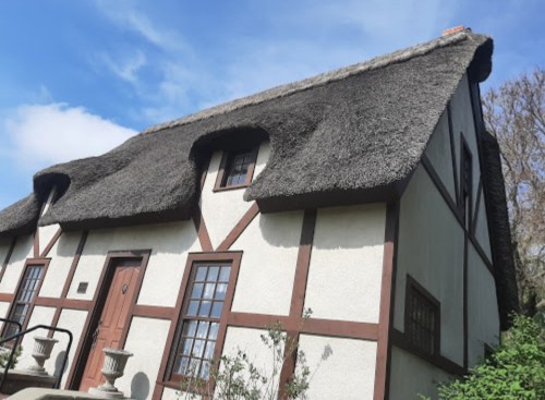 A charming thatched-roof cottage with white walls and wooden beams, surrounded by greenery under a blue sky.