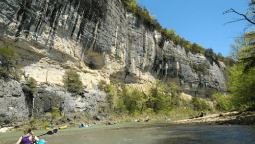 A scenic river with kayakers paddling alongside towering cliffs and lush greenery under a clear blue sky.