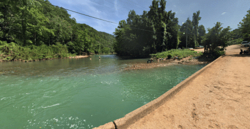 A serene river scene surrounded by lush greenery, with people enjoying the water and a dirt path along the bank.