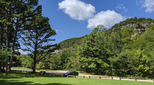 A scenic view of green trees and a rocky cliff under a blue sky with fluffy clouds, featuring a parked car.