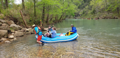 A group of four people in a blue raft at a riverbank, surrounded by trees and rocky terrain.