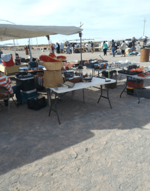 A bustling outdoor market with tables displaying various goods and people browsing in the background.