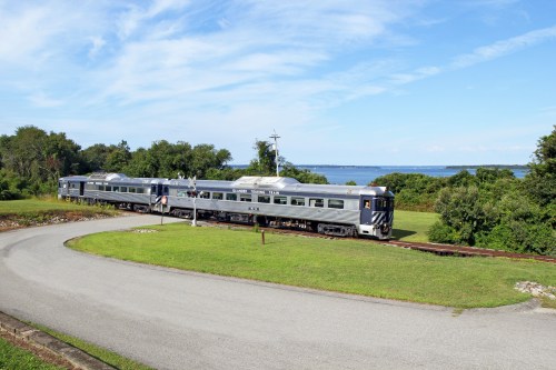 A vintage train travels along a scenic route near a body of water, surrounded by lush greenery and blue skies.