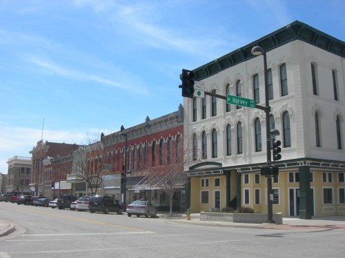 Historic buildings line a street with traffic lights, blue sky, and parked cars in a small town.