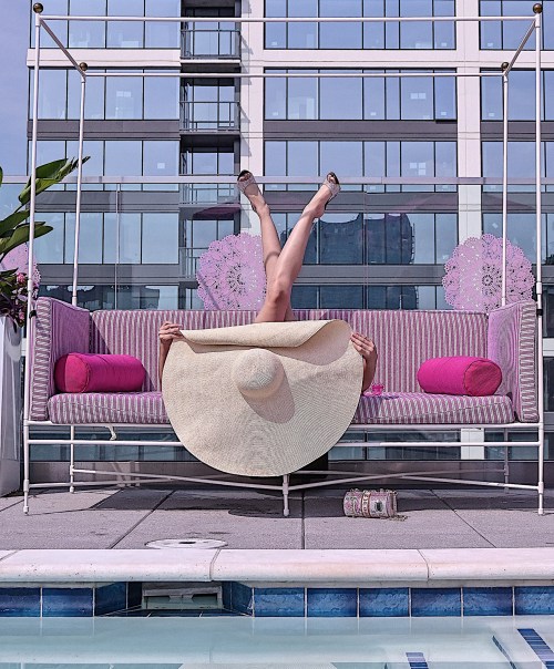A person lounges on a pink-striped sofa by a pool, wearing a large sun hat and showing their legs in the air.