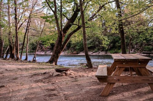 A peaceful riverside scene featuring a wooden picnic table, trees, and a calm water view.