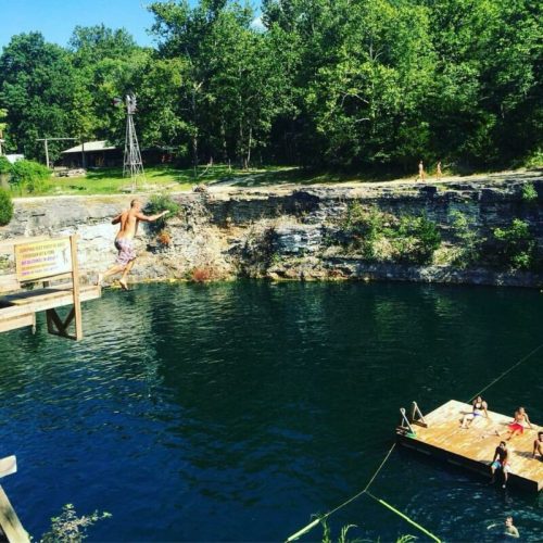 A person jumps off a wooden platform into a deep blue swimming hole, surrounded by trees and a small dock with people.