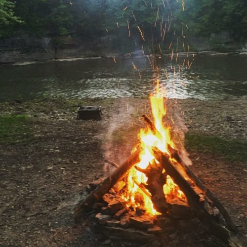 A campfire crackles by a riverbank, with sparks flying into the air amidst a serene, wooded backdrop.