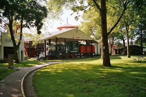 A vintage steam locomotive displayed under a shelter, surrounded by trees and green grass in a park setting.