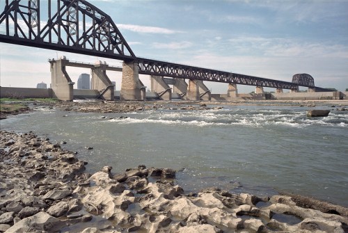 A river flows beneath a large metal bridge, with rocky banks and a city skyline in the background on a clear day.