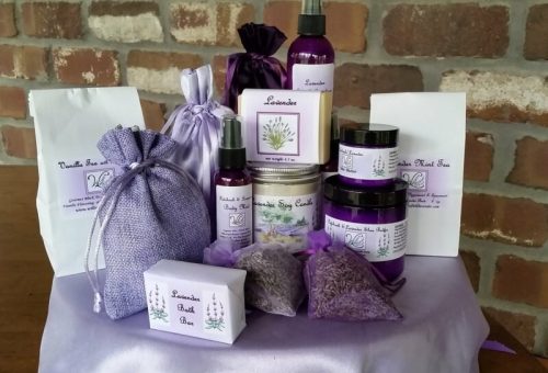 A collection of lavender-themed products, including sprays, soaps, and sachets, arranged on a table with a brick background.