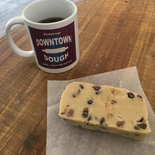 A mug of coffee next to a piece of edible cookie dough on a wooden table.