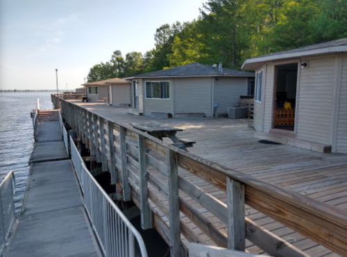 A wooden dock extends over water, with cabins lined along the side and trees in the background.
