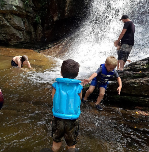 Children playing in a waterfall pool, one wearing a blue life jacket, while adults supervise nearby.