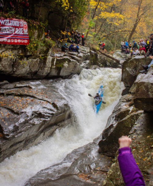 A kayaker navigates a rapid in a rocky river while spectators watch from the banks, surrounded by autumn foliage.