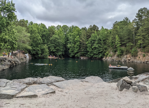 A serene lake surrounded by lush green trees, with people swimming and a small boat on the water.
