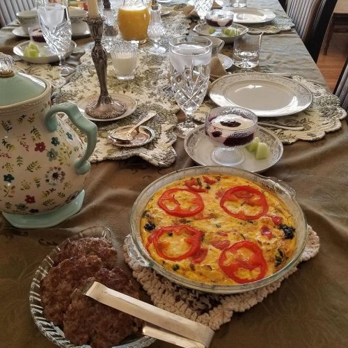 A beautifully set dining table with a frittata, sausage, and drinks, surrounded by elegant glassware and tableware.