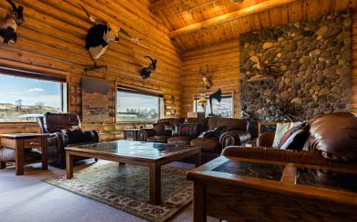 Cozy log cabin interior with leather furniture, stone fireplace, and large windows showcasing a scenic view.