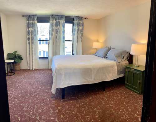 A cozy bedroom featuring a bed, two lamps, a green nightstand, and large windows with patterned curtains.