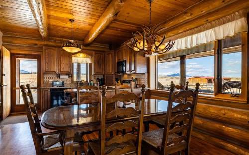 Cozy log cabin kitchen with wooden furniture, large windows, and rustic decor, featuring a dining table and chandelier.