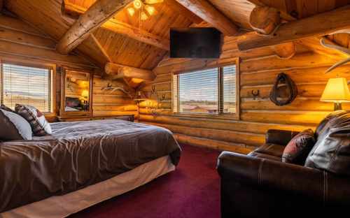 Cozy log cabin bedroom with wooden beams, a large bed, a TV, and a seating area by the window. Warm, inviting atmosphere.