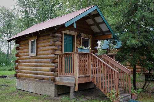 A small log cabin with a green door and a wooden porch, surrounded by trees and greenery.
