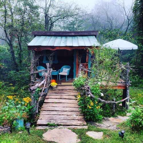 A cozy cabin surrounded by greenery and flowers, with a wooden walkway and blue chairs on a misty day.