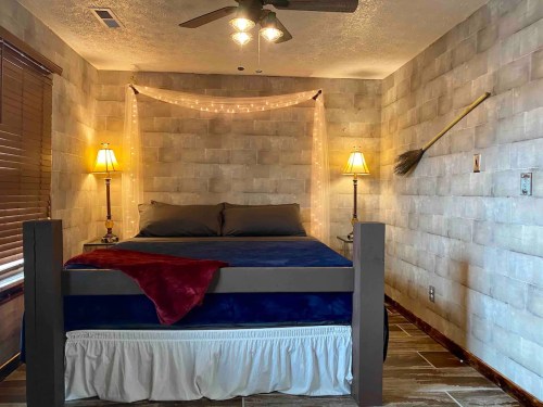Cozy bedroom with a bed, decorative lights, lamps, and a broom on the wall, featuring a rustic stone wall design.