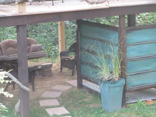A cozy outdoor seating area with a wooden structure, green walls, and potted plants surrounded by greenery.
