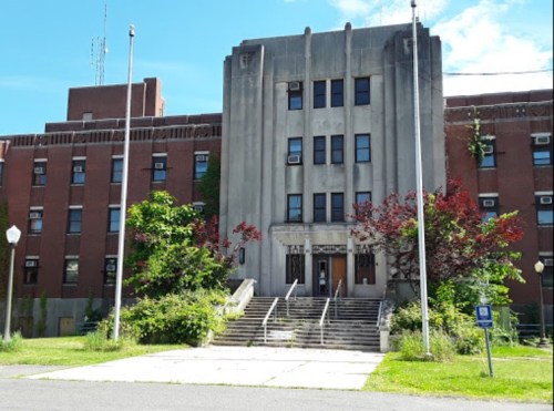 Historic building with a grand entrance, surrounded by overgrown greenery and a clear blue sky.