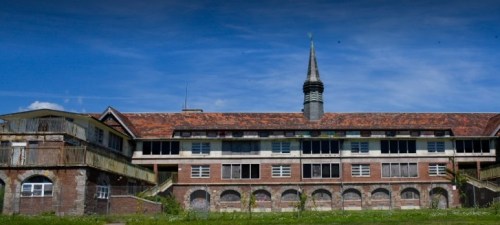 Abandoned building with a steeple, featuring brick walls and overgrown grass under a blue sky.