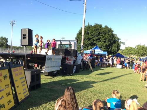 Children perform on a stage at an outdoor event, with a crowd and vendor tents in the background.