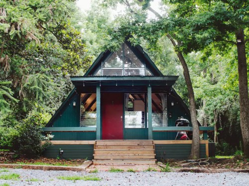 A charming A-frame cabin surrounded by trees, featuring a red door and large windows.