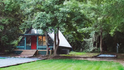 A modern A-frame cabin surrounded by lush greenery and trees, with a red door and a gravel pathway.