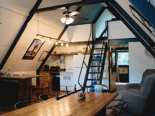 Cozy A-frame cabin interior with a kitchen, dining table, and lofted sleeping area, featuring modern decor and warm lighting.