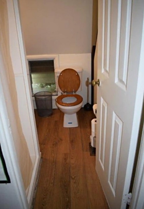 A small bathroom with a wooden toilet seat, a wastebasket, and a door leading to another room.