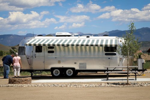 A shiny silver Airstream trailer with a striped awning, parked near a picnic table and surrounded by mountains.