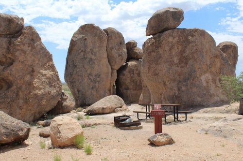 Rock formations surround a picnic area with a table and grill under a blue sky with scattered clouds.