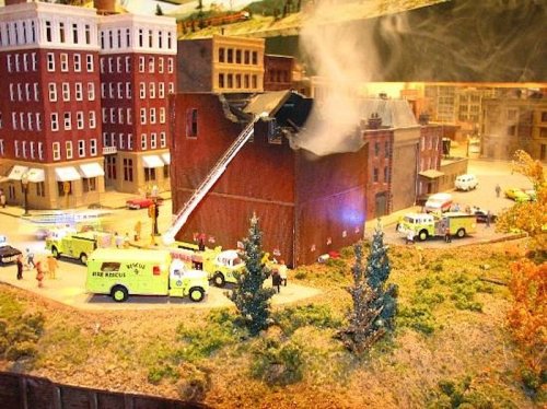 A miniature fire scene with toy fire trucks, firefighters, and smoke rising from a model building in a detailed diorama.