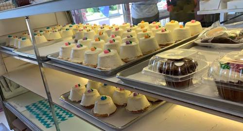 A display of small, frosted cakes with colorful decorations on top, arranged on trays in a bakery.
