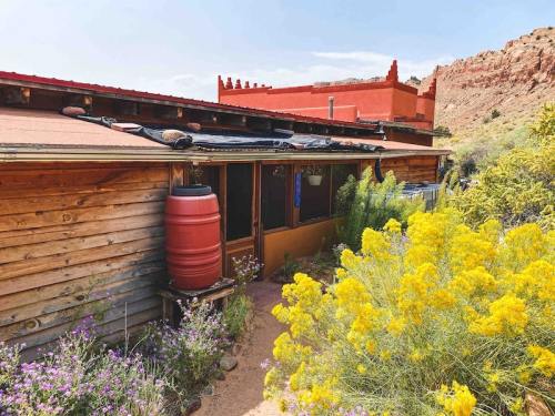 A rustic wooden building surrounded by vibrant yellow flowers and a red water barrel, set against rocky terrain.