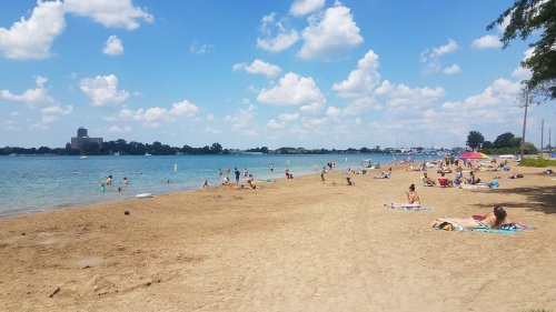 A sunny beach scene with people swimming, sunbathing, and enjoying the water under a blue sky with fluffy clouds.