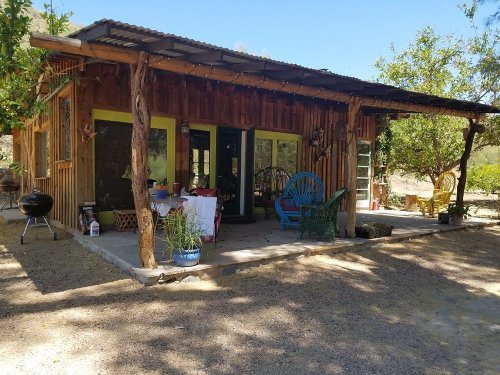 A rustic cabin with a porch, surrounded by trees and outdoor seating, set in a sunny, natural landscape.