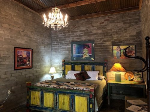 Cozy bedroom with a colorful wooden bed, chandelier, and artwork on stone walls, creating a warm, inviting atmosphere.