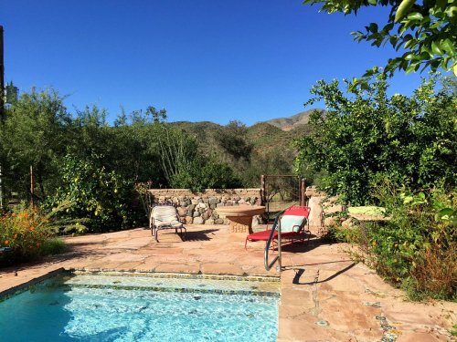 A serene outdoor space featuring a pool, lounge chairs, and lush greenery with mountains in the background under a clear blue sky.