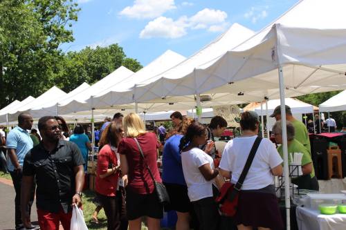 A bustling outdoor market with people browsing under white tents on a sunny day.