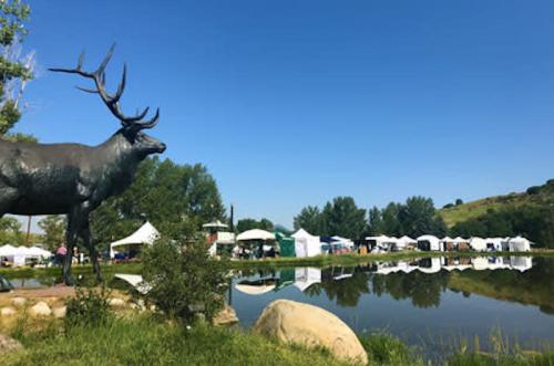 A bronze elk statue stands by a pond, with white tents and trees in the background under a clear blue sky.
