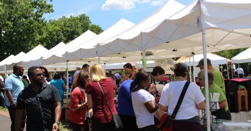 A bustling outdoor market with people browsing under white tents on a sunny day.
