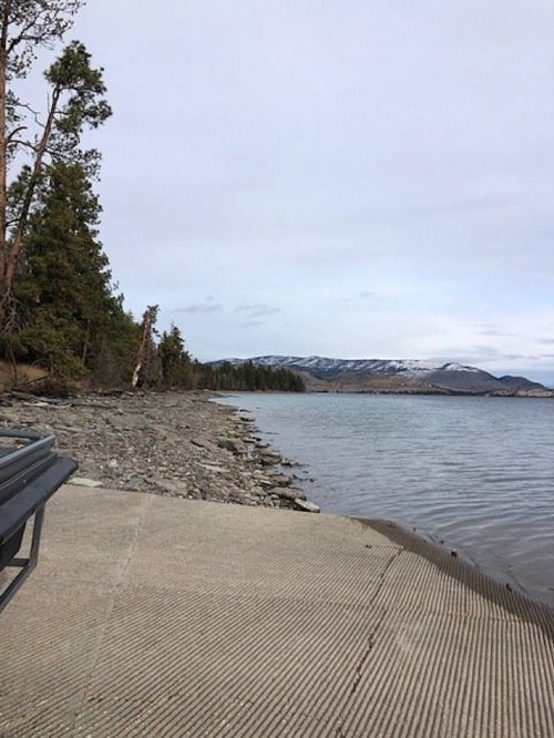 A serene lakeside view with rocky shore, trees, and distant mountains under a cloudy sky.